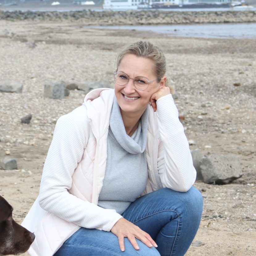 Eine Frau sitzt am Strand mit einem Hund und lächelt in die Kamera.